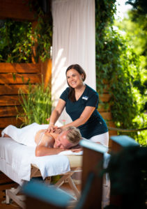 Eine Wellness-Mitarbeiterin führt eine Massage an einer liegenden Person durch. Die Mitarbeiterin trägt ein schwarzes T-Shirt und weiße Hosen.
