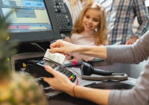 Eine Kassenmitarbeiterin scannt eine Karte an einem Kassenterminal. Im Hintergrund sind Kunden zu sehen. Auf dem Bildschirm sind Zahlen und eine Benutzeroberfläche sichtbar.