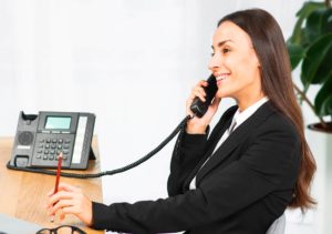 Eine Kassenmitarbeiterin trägt einen schwarzen Anzug und hält einen Telefonhörer in der linken Hand. Auf dem Tisch liegt ein Stift.