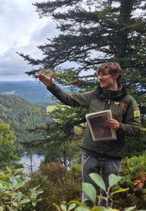 Person in Outdoorjacke hält eine Broschüre und zeigt mit der Hand auf eine bewaldete Landschaft mit See im Hintergrund.