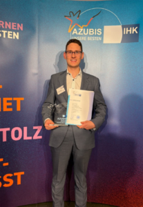 Mann im Anzug hält Urkunde und Trophäe vor blauem Hintergrund mit Schriftzug 'AZUBIS UNSERE BESTEN IHK'